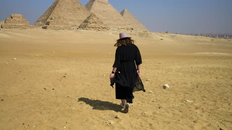 Sandy-Ground-With-Girl-Wearing-Black-Dress-Walking-Towards-Great-Pyramids-Of-Giza-In-Cairo,-Egypt