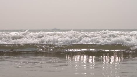 Close-up-of-Pacific-ocean-waves-crashing-down-onto-the-sandy-beach-San-Diego,-California-with-a-large-ship-in-the-background