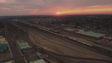 Zooming-out-cinematic-top-city-aerial-view-of-downtown-Roseville,-California-with-industrial-buildings,-shops-and-cargo-train-passing-down-the-railroad-with-a-vivid-orange-pink-sunset-in-background