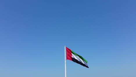 Vista-Aérea-De-La-Bandera-De-Los-Emiratos-árabes-Unidos-Levantándose-En-Un-Hermoso-Día-Soleado,-Cielo-Azul-En-El-Fondo,-El-Símbolo-Nacional-De-Los-Emiratos-árabes-Unidos