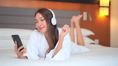 Feliz-Mujer-Asiática-Bonita-Mirando-El-Teléfono-Y-Escuchando-Música-Mientras-Está-Acostada-En-La-Cama-Del-Hotel-Usando-Una-Bata-Blanca,-Vista-Frontal-Sonriente
