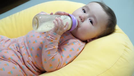 Caucasian-baby-girl-holding-bottle-and-drinking-formula-milk-while-Lying-On-A-Yellow-Pillow--super-close-up