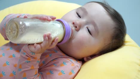 Baby-girl-holding-bottle-with-two-hands-and-drinking-formula-milk-while-Lying-On-A-Yellow-Pillow--super-close-up