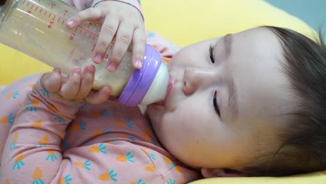 Infant-nine-month-baby-girl-holding-bottle-with-formula-milk-and-drinking-by-herself-Lying-Down-On-A-Yellow-Pillow---close-up