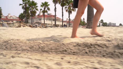Tiefschuss-Von-Weiblichen-Beinen,-Die-Auf-Goldenem-Sand-Mit-Palmen-Und-Resort-Im-Hintergrund-Zum-Strand-Hinuntergehen