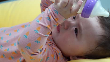 Baby-Girl-Holding-And-Drinking-Milk-From-A-Glass-Feeding-Bottle-While-Lying-Down-On-A-Yellow-Pillow---close-up