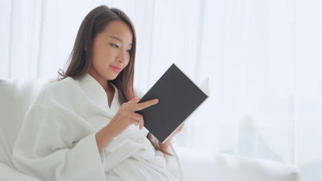 Smiling-pretty-Asian-woman-lying-on-the-hotel-white-bed-wearing-a-white-dressing-gown-and-reading-a-book-in-the-morning-daytime