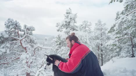 Man-Seats-On-Winter-Valley-While-Embracing-His-Alaskan-Malamute-Dog-During-Snowy-Day-Near-Trondheim,-Norway