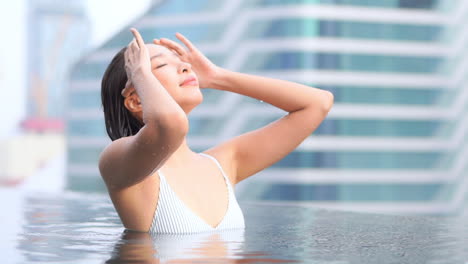 Hübsche-Asiatische-Frau-In-Einem-Schwimmbad,-Die-Ihr-Nasses-Haar-Streichelt,-Mit-Stadtgebäuden-Im-Hintergrund,-Zeitlupe
