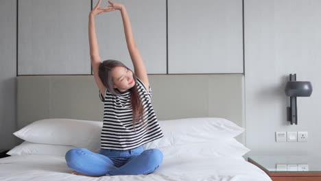 Pretty-young-smiled-Asian-woman-stretching-her-hands-up-while-sitting-on-a-bed-with-crossed-legs-in-the-morning