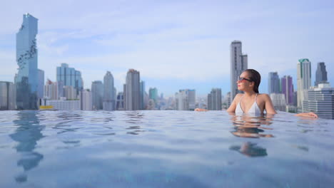 Attractive-asian-woman-in-infinity-pool-with-stunning-view-of-Bangkok,-Thailand-cityscape,-luxury-vacation-and-fashion-concept