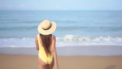 Young-Asian-woman-wearing-yellow-swimming-suit-going-to-swim,-walking-towards-the-sea-at-the-picturesque-island,-slow-motion-back-view