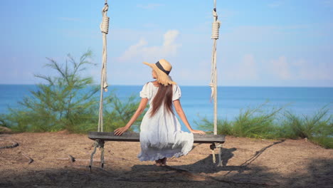 Young-woman-sitting-and-swinging-on-a-wooden-swing-hanging-on-a-tree-at-a-tropical-beach-in-Thailand-enjoying-the-sea-view,-slow-motion