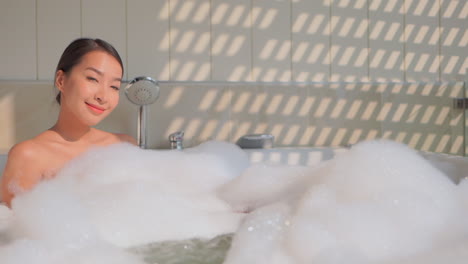 Woman-taking-a-hot-bath-relaxing-in-a-bathtub-filled-with-foam-outside-under-sunlight,-Asian-female-looking-into-camera-and-smiles