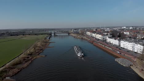 Following-a-large-container-cargo-ship-passing-by-embankment-of-historic-medieval-Hanseatic-city-of-Zutphen,-The-Netherlands,-besides-river-IJssel