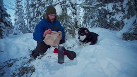 Man-With-His-Alaskan-Malamute-Pet-Dog-Sitting-On-Snowy-Ground-And-Having-Snack-During-Work-Break-In-Trondheim,-Norway---static-shot
