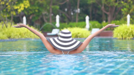 Back-view-of-woman-in-black-and-white-hat-raising-hands-from-water-in-swimming-pool-on-blurred-greenery-background-daytime-slow-motion