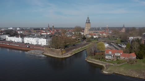 Picturesque-aerial-rotate-showing-medieval-Hanseatic-city-Zutphen,-The-Netherlands,-with-entrance-to-small-Vispoorthaven-or-Gelre-port-and-cargo-ship-passing-on-river-IJssel-against-a-blue-sky