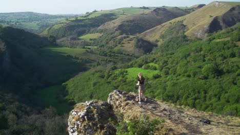 Slow-mo-of-young-blonde-woman-walking-towards-edge-of-cliff-above-Thor's-cave,-Ashbourne,-Peak-District,-England-while-flicking-long-hair-back-over-shoulders