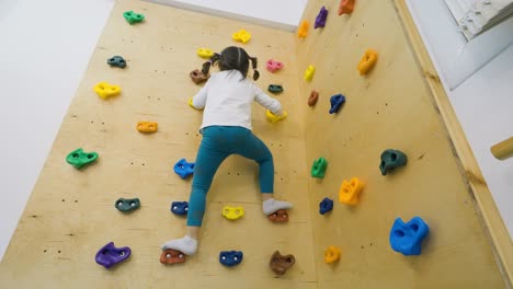 Wide-shot-of-a-fearless-little-girl-climbing-an-indoor-rock-climbing-wall