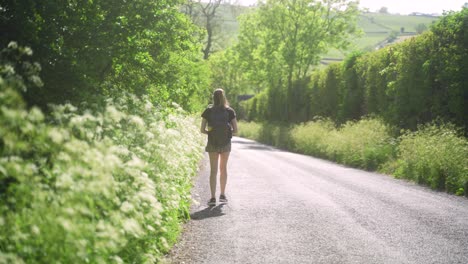 Handheld-shot-of-blonde-young-woman-walking-down-a-British-country-road,-Peak-District,-England,-lined-with-overgrown-cows-parsley-and-green-hedges-wearing-a-backpack