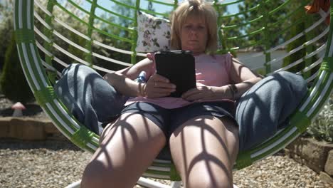 Woman-relaxes-in-comfortable-garden-swing-chair-reading-digital-eBook-on-warm-sunny-day