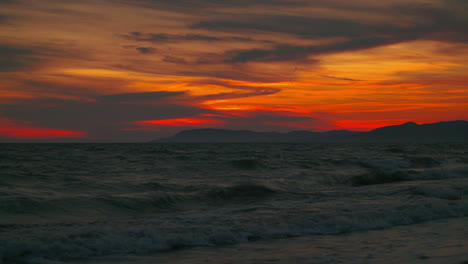 4K-UHD-Cinemagraph-seamless-video-loop-of-of-the-sunset-seen-from-a-romantic-sandy-beach-with-mountains-and-waves-at-the-beautiful-Italian-Mediterranean-seaside-with-red-and-orange-clouds-in-the-sky