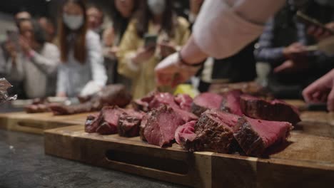 Chef-puts-freshly-grilled-meat-slices-together-on-wooden-cut-board-at-meat-tasting-event