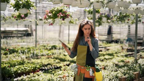 Red-haired-woman-gardener-with-tablet-walking-in-a-greenhouse-with-plants