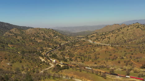 A-long-serpentine-train-winds-through-the-Tehachapi-foothills-below-the-highway-traffic---aerial-view