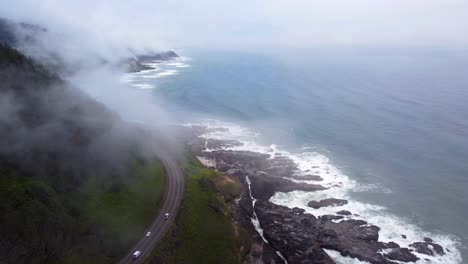 4K-30FPS-Aerial-Footage-Oregon-Coast---Epic-Static-Shot-of-US-Route-101-and-car-traffic--Flying-amongst-clouds,-waves-crashing-against-mossy-stone-rocks-on-the-Pacific-Ocean-Shore---DJI-Drone-Video