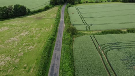 Eine-Langsam-Absteigende-Und-Aufsteigende-Kameraaufnahme,-Die-Ein-Auto-Zeigt,-Das-An-Einem-Bewölkten-Tag-In-England-Einen-Schmalen-Landweg-Zwischen-Zwei-Großen-Feldern-Hinunterrast