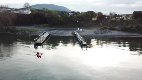 Aerial-View,-a-man-kayaking-out-to-the-lake-from-a-jetty,-in-the-early-springtime-on-a-calm-day-in-New-Zealand,-Pan-Slowly