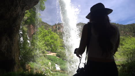 Back-of-female-walking-in-cave-under-scenic-waterfall-on-sunny-summer-day