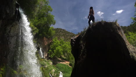 Young-Woman-Standing-on-Rock-in-Front-of-Amazing-Waterfalls,-Low-Angle-Cinematic-View