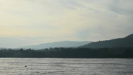 A-boat-crossing-the-Mekong-river-in-a-diagonal-direction-towards-Laos-from-Thailand-with-a-background-of-forest-and-hills