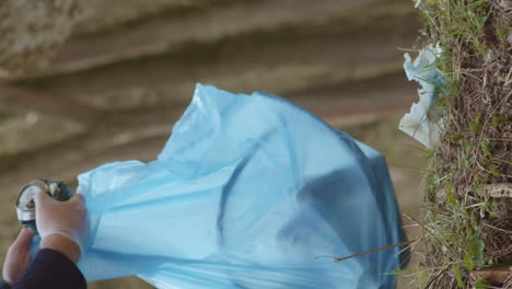 Man-in-gloves-gathers-litter-in-bag-in-woods,-vertical,-slow-motion