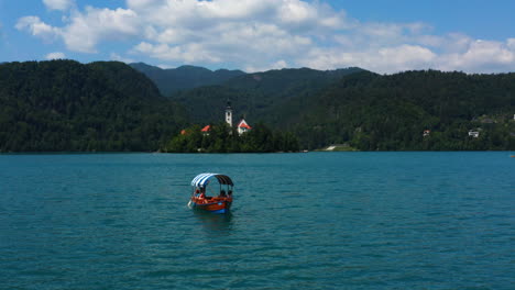 Touristen,-Die-Auf-Einem-Pletna-boot-Den-See-Kreuzen,-Mit-Blick-Auf-Die-Kirche-Der-Königin-Maria-Und-Die-Bergkette-In-Slowenien