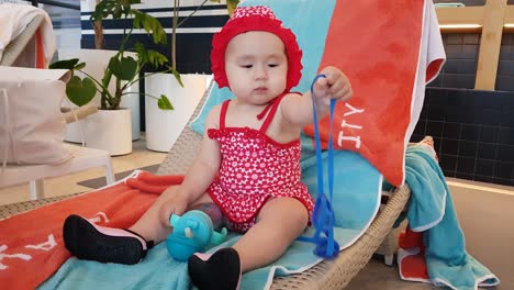 Cute-Baby-Girl-Holding-Blue-Strap-And-Water-Tumbler-On-Lounge-Chair-By-The-Poolside