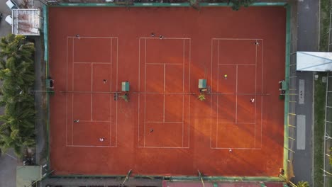 Aerial-view-in-slow-motion-of-clay-tennis-courts,-with-people-playing-wearing-white-clothes