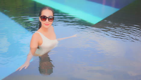 Schöne-Asiatische-Frau-Im-Futuristischen-Luxus-Swimmingpool-Mit-Sonnenbrille-Und-Weißem-One-Shoulder-Monokini,-Die-Auf-Die-Kameralinse-Blickt-Und-Sich-Am-Rand-Des-Pools-Lehnt