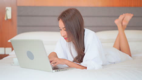 Woman-in-bathrobe-typing-on-laptop-computer-keyboard-while-lying-on-the-bed-in-a-luxury-hotel
