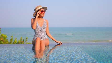 A-pretty-young-woman-sits-in-the-shallow-water-on-the-edge-of-a-swimming-pool-with-the-ocean-horizon-in-the-background