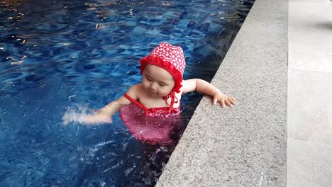 Verspieltes-Babymädchen-Im-Badeanzug-Steht-Auf-Der-Stufe-Des-Schwimmbeckens-Und-Spielt-Und-Schlägt-Wasser-Mit-Der-Hand