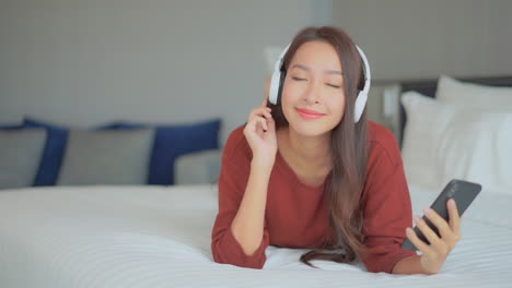 Happy-pretty-Asian-woman-watching-phone-and-listen-to-music-while-lying-on-the-hotel-bed-wearing-casual-clothes,-smiling-front-view