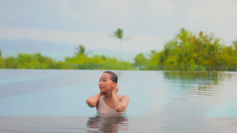 Attractive-young-woman-touches-her-wet-hair-inside-the-swimming-pool-water-of-an-exotic-Bali-hotel-with-closed-eyes-on-a-sunny-day,-portrait-slow-motion