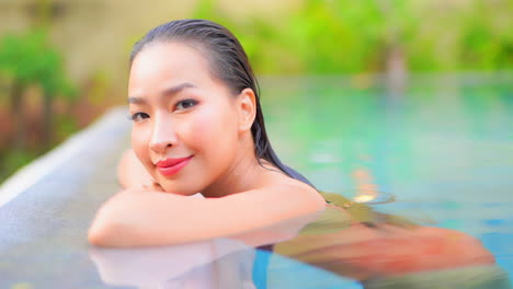 Primer-Plano-De-La-Cara-De-Una-Mujer-Asiática-Con-Una-Piel-Perfecta-Apoyada-En-Los-Brazos-Y-Sonriendo-En-El-Borde-De-La-Piscina-En-El-Resort-Tópico-En-El-Día-De-Bali