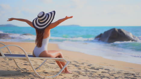Unrecognizable-fit-girl-in-swimsuit-and-striped-hat-sitting-on-deckchair-near-the-sea-when-water-rolling-over-sandy-beach-daytime