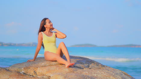 Beautiful-Asian-woman-in-yellow-monokini-sitting-on-a-big-rock-on-the-seafront-at-Philippine's-island-and-touching-her-hair,-slow-motion-handheld
