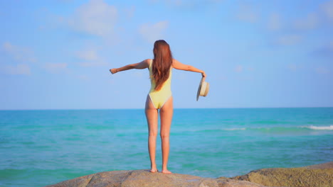 Back-to-the-camera,-a-healthy,-fit-woman-in-a-bathing-suit-stands-on-a-huge-rock-facing-the-ocean-horizon